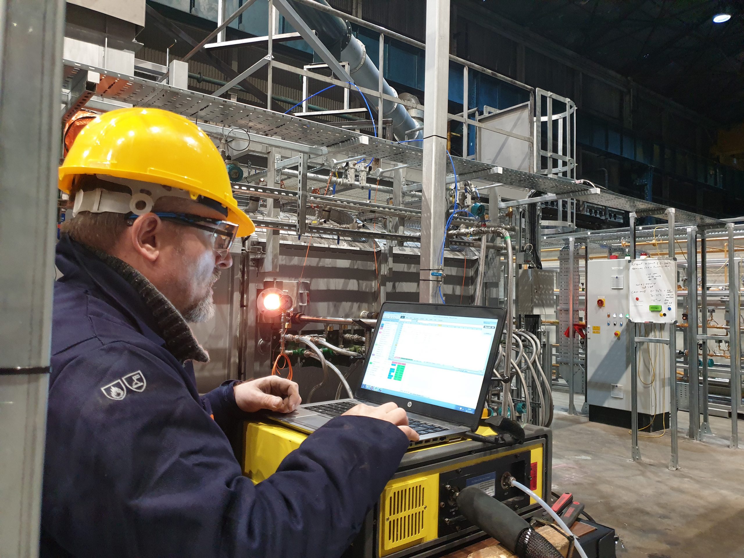 Kris Milkowski from TERC wears a hard hat and works at a laptop inside a building with lots of metal pipework