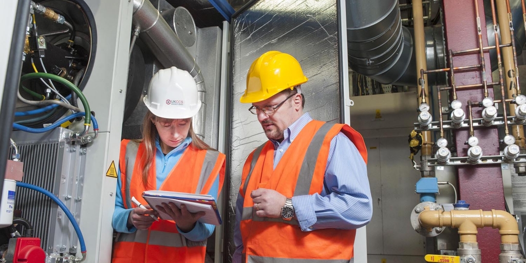 drp5369-resized a man and a woman look at a notepad, stood next to a piece of research equipment, both are wearing safety hats and hi-vis jackets
