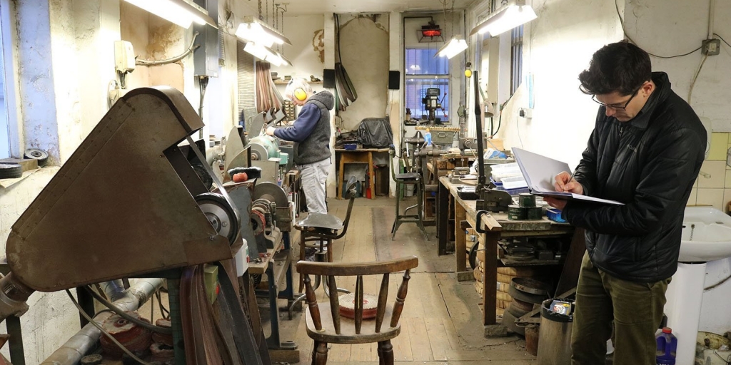 img_2047-resized a man writes on a clipboard in the foreground of a metalwork workshop, in the background a man in ear defenders works on equipment